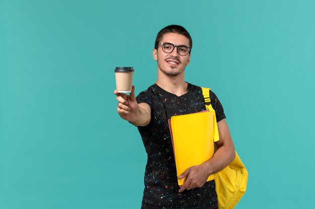 Vorderansicht des männlichen Studenten im gelben Rucksack des dunklen T-Shirts, der verschiedene Dateien und Kaffee an der blauen Wand hält