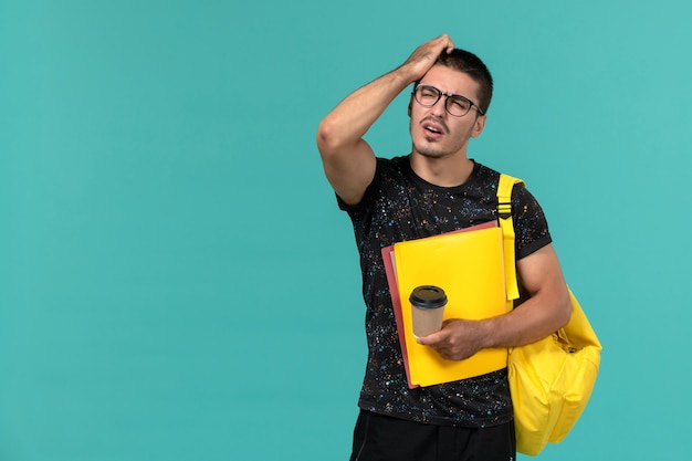 Vorderansicht des männlichen Studenten im gelben Rucksack des dunklen T-Shirts, der verschiedene Dateien und Kaffee denkt, die auf blaue Wand denken