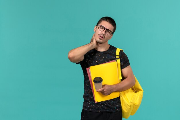 Vorderansicht des männlichen Studenten im gelben Rucksack des dunklen T-Shirts, der verschiedene Dateien und Kaffee hält, die Nackenschmerzen auf blauer Wand haben