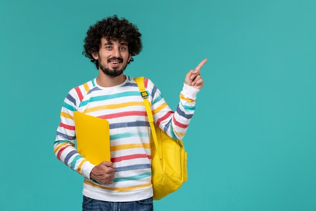 Vorderansicht des männlichen Studenten im gestreiften Hemd, das gelben Rucksack hält, der Dateien hält, die auf der blauen Wand lächeln