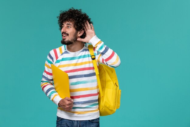 Vorderansicht des männlichen Studenten im gestreiften Hemd, das gelben Rucksack hält, der Dateien hält, die versuchen, auf blauer Wand zu hören