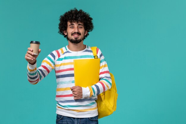 Vorderansicht des männlichen Studenten im gestreiften Hemd, das gelben Rucksack hält, der Dateien und Kaffee auf blauer Wand hält