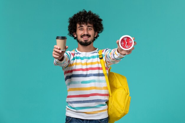 Vorderansicht des männlichen Studenten im gestreiften Hemd, das gelben Rucksack hält, der Kaffee und Uhren an der blauen Wand hält