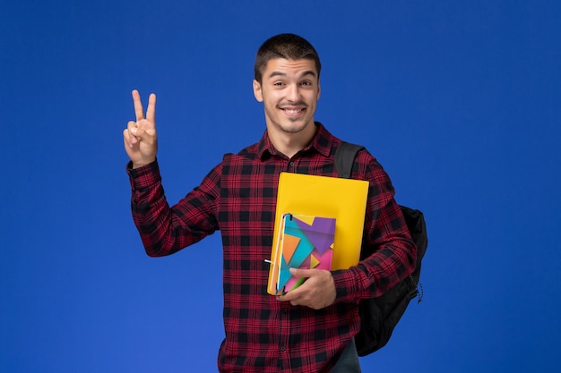 Vorderansicht des männlichen Studenten im roten karierten Hemd mit dem Rucksack, der Dateien und die lächelnden Hefte hält