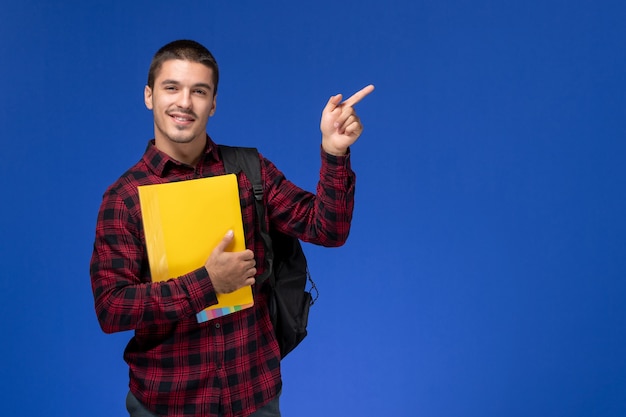 Vorderansicht des männlichen Studenten im roten karierten Hemd mit dem Rucksack, der gelbe Dateien hält, die auf der blauen Wand lächeln
