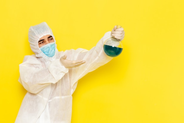 Vorderansicht des männlichen wissenschaftlichen Arbeiters im speziellen Schutzanzug, der Flasche mit grüner Lösung auf gelber Wand hält