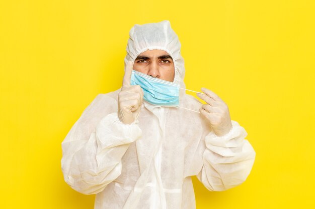 Vorderansicht des männlichen wissenschaftlichen Arbeiters im speziellen Schutzanzug, der seine Maske auf gelber Wand abnimmt