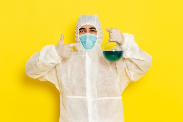 Vorderansicht des männlichen wissenschaftlichen Arbeiters im speziellen Schutzanzug mit der Maskenhalteflasche auf gelbem Schreibtisch-Wissenschaftsarbeiter-Kostümfarbfoto