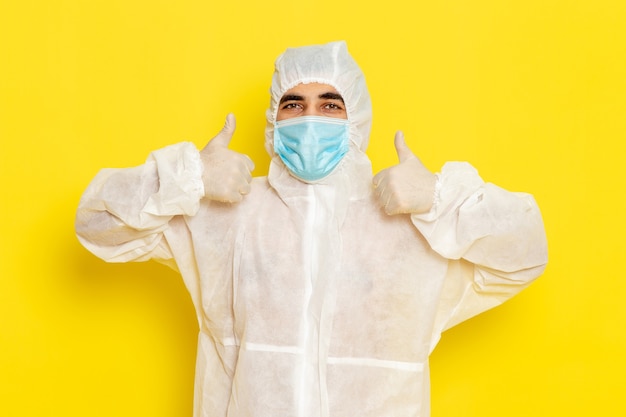 Vorderansicht des männlichen wissenschaftlichen Arbeiters im speziellen weißen Schutzanzug mit der Maske, die auf gelber Wand lächelt