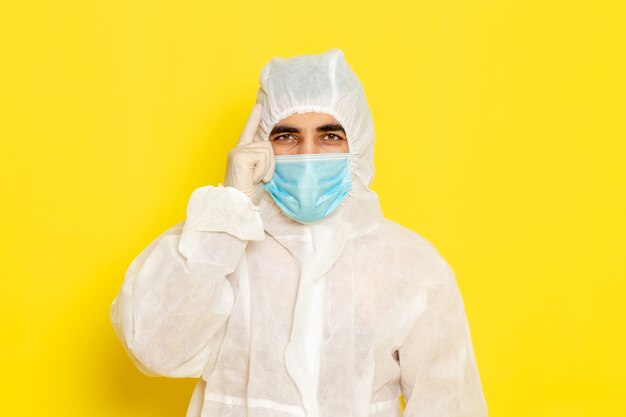 Vorderansicht des männlichen wissenschaftlichen Arbeiters im speziellen weißen Schutzanzug mit Maske an der hellgelben Wand