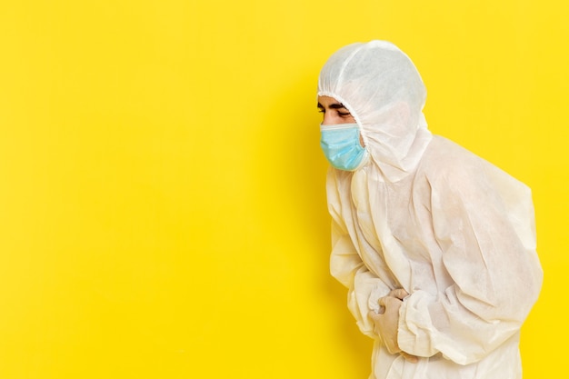 Vorderansicht des männlichen wissenschaftlichen Arbeiters im speziellen weißen Schutzanzug und mit Maske, die seinen Magen auf hellgelbem Schreibtisch-Wissenschaftsarbeiterchemie-Farbgefahrfoto hält