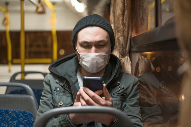 Vorderansicht des Mannes mit der medizinischen Maske im Bus, der sein Telefon betrachtet