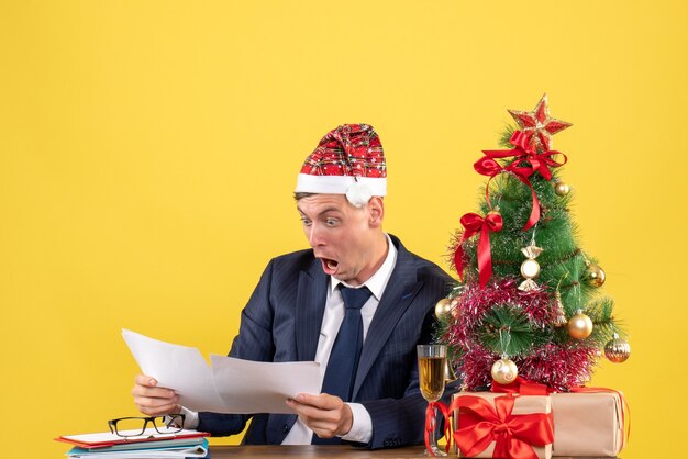 Vorderansicht des Mannes mit großen Augen, der Papiere prüft, die am Tisch nahe Weihnachtsbaum sitzen und auf Gelb präsentieren