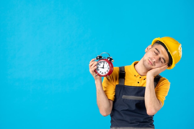 Vorderansicht des müden männlichen Arbeiters in der gelben Uniform mit den Uhren auf Blau