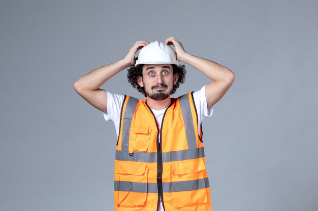 Vorderansicht des nachdenklichen männlichen Baumeisters, der Warnweste mit Schutzhelm auf grauer Wellenwand trägt