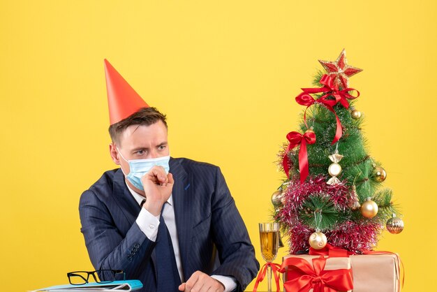 Vorderansicht des nachdenklichen Mannes mit der medizinischen Maske, die am Tisch nahe Weihnachtsbaum sitzt und auf Gelb präsentiert