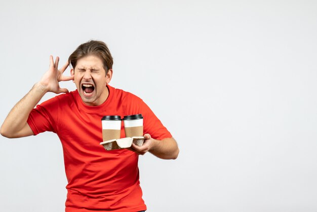 Vorderansicht des nervösen jungen Mannes in der roten Bluse, die Kaffee in Pappbechern hält und eines seiner Ohren auf weißem Hintergrund schließt