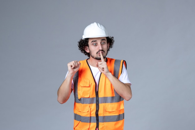 Vorderansicht des nervösen männlichen Baumeisters, der eine Warnweste mit Schutzhelm trägt und auf der grauen Wellenwand eine stille Geste macht