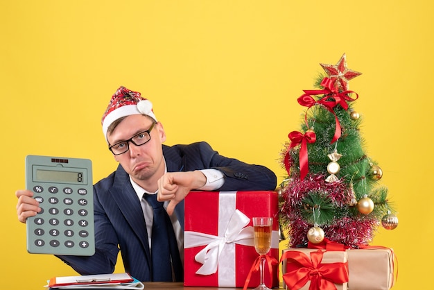 Vorderansicht des niedergedrückten Geschäftsmannes mit Weihnachtsmütze, die am Tisch nahe Weihnachtsbaum sitzt und auf Gelb präsentiert