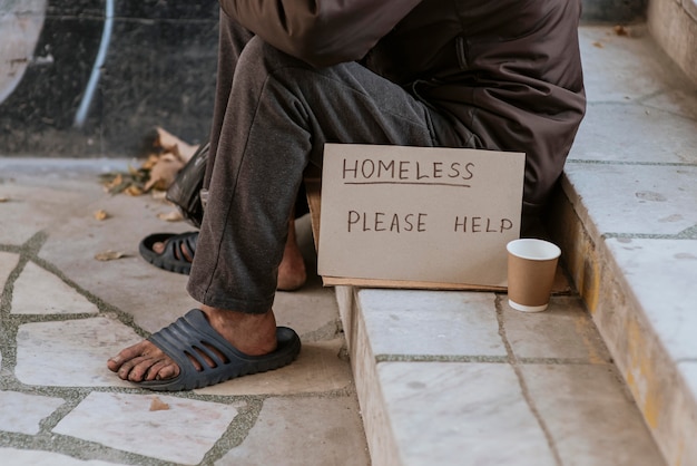 Vorderansicht des Obdachlosen auf der Treppe mit Hilfeschild und Tasse