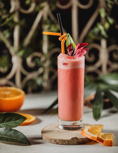 Vorderansicht des roten Grapefruitsmoothie in einem Glas mit Strohhalm und geschnittenen Orangen auf einem hölzernen Ständer auf dem Tisch