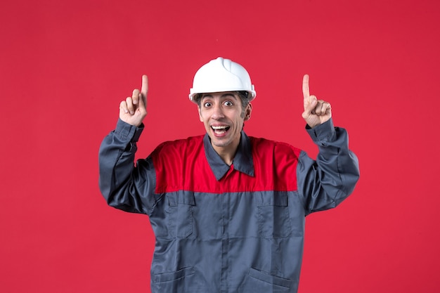 Vorderansicht des schockierten jungen Baumeisters in Uniform mit Schutzhelm, der auf isolierte rote Wand zeigt