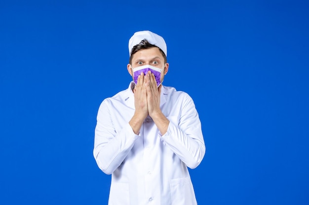 Vorderansicht des schockierten männlichen Arztes im medizinischen Anzug und in der lila Maske auf Blau