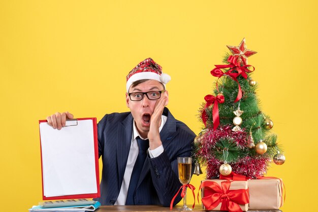 Vorderansicht des schockierten Mannes, der Klemmbrett hält, das am Tisch nahe Weihnachtsbaum sitzt und auf Gelb präsentiert