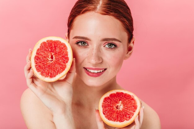 Vorderansicht des schönen kaukasischen Mädchens mit Grapefruit Studioaufnahme einer positiven Ingwerfrau mit geschnittenen Zitrusfrüchten