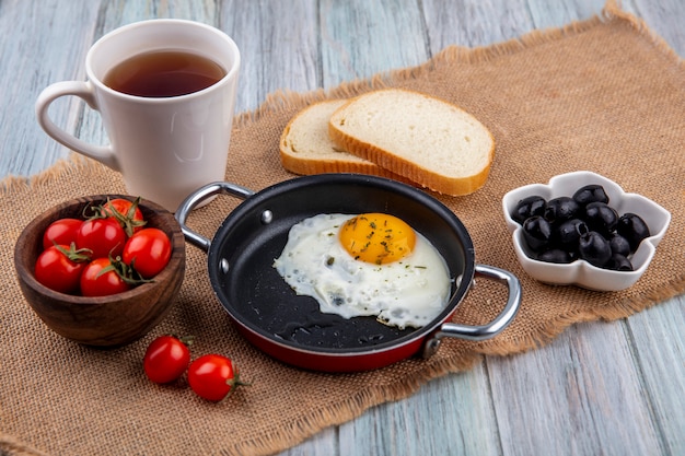 Vorderansicht des Spiegeleis in der Pfanne mit Schüssel Tomate und Tee mit Brotscheiben und Schüssel Olive auf Sackleinen und Holzoberfläche