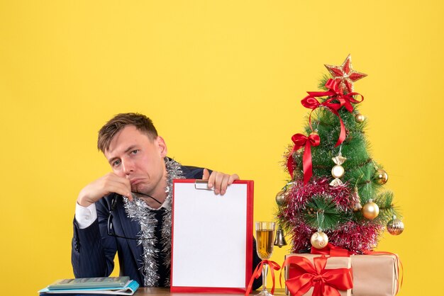 Vorderansicht des traurigen Geschäftsmannes, der am Tisch nahe Weihnachtsbaum sitzt und auf Gelb präsentiert