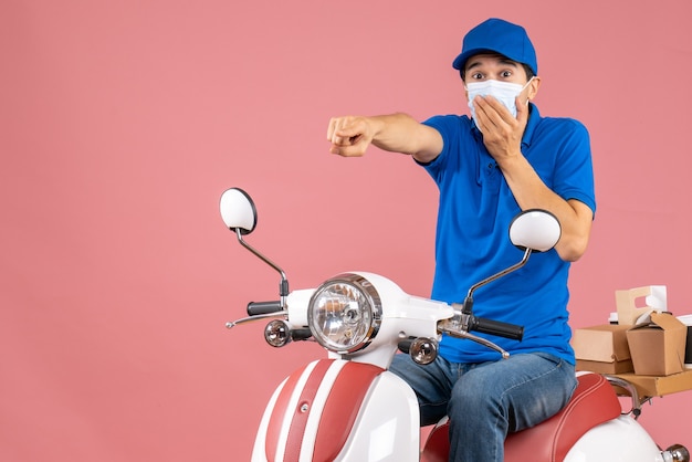 Vorderansicht des überraschten Lieferers in medizinischer Maske mit Hut, der auf einem Roller sitzt und nach vorne auf pastellfarbenem Pfirsichhintergrund zeigt