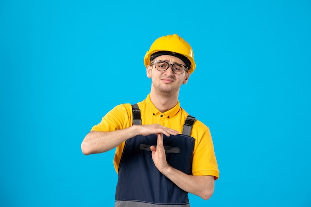 Vorderansicht des unzufriedenen männlichen Arbeiters in der gelben Uniform, die t Zeichen auf blauer Wand zeigt