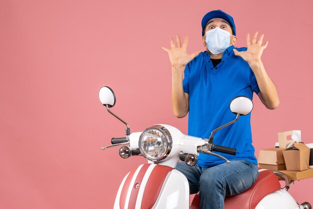 Vorderansicht des verängstigten Lieferboten in medizinischer Maske mit Hut, der auf einem Roller auf pastellfarbenem Pfirsichhintergrund sitzt