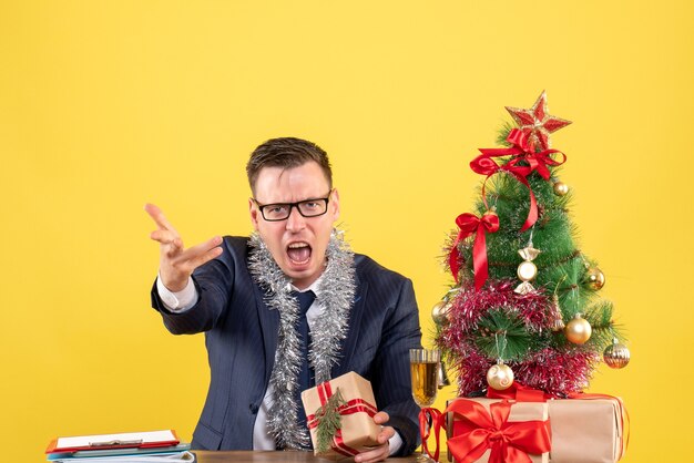 Vorderansicht des verärgerten Mannes mit Brille, die am Tisch nahe Weihnachtsbaum sitzt und auf gelber Wand präsentiert