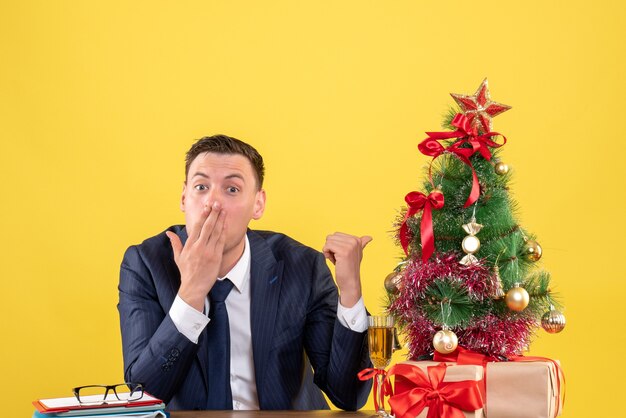 Vorderansicht des verblüfften Mannfingers, der Weihnachtsbaum zeigt, der am Tisch nahe Weihnachtsbaum und Geschenke auf gelber Wand sitzt