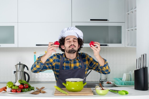 Vorderansicht des verträumten männlichen Kochs mit frischem Gemüse, das rote Paprika in der weißen Küche hält