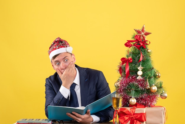 Vorderansicht des verwirrten Geschäftsmannes, der am Tisch nahe Weihnachtsbaum sitzt und auf Gelb präsentiert