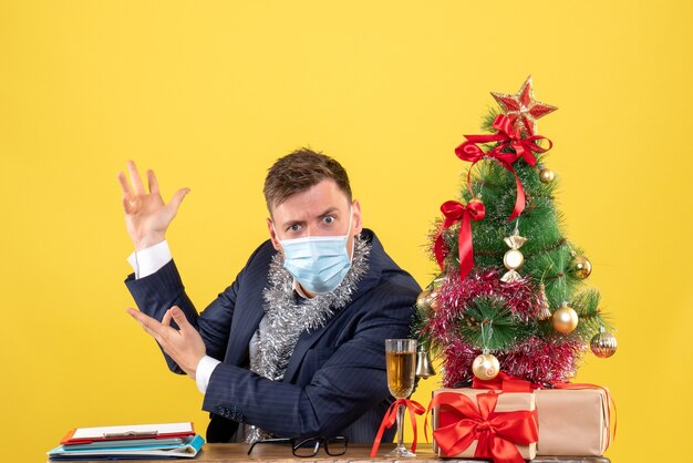 Vorderansicht des verwirrten Geschäftsmannes, der am Tisch nahe Weihnachtsbaum sitzt und auf Gelb präsentiert