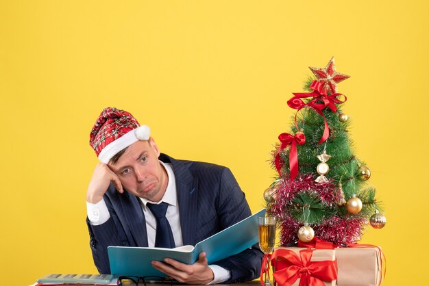 Vorderansicht des verwirrten Geschäftsmannes, der am Tisch nahe Weihnachtsbaum sitzt und auf Gelb präsentiert