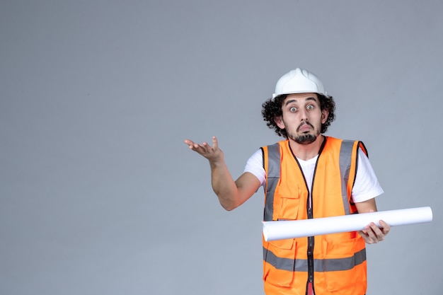 Vorderansicht des verwirrten männlichen Konstrukteurs in Warnweste mit Schutzhelm und leerer grauer Wand