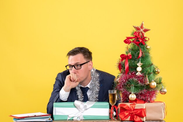 Vorderansicht des verwirrten Mannes, der am Tisch nahe Weihnachtsbaum sitzt und auf Gelb präsentiert