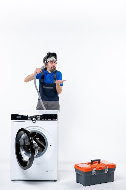 Kostenloses Foto vorderansicht des verwirrten mechanikers in uniform, der hinter der waschmaschine steht und das abflussrohr an der weißen wand ausbläst