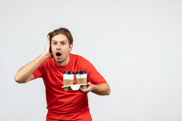 Vorderansicht des verwirrten und emotionalen jungen Kerls in der roten Bluse, die Kaffee in den Pappbechern auf weißem Hintergrund hält