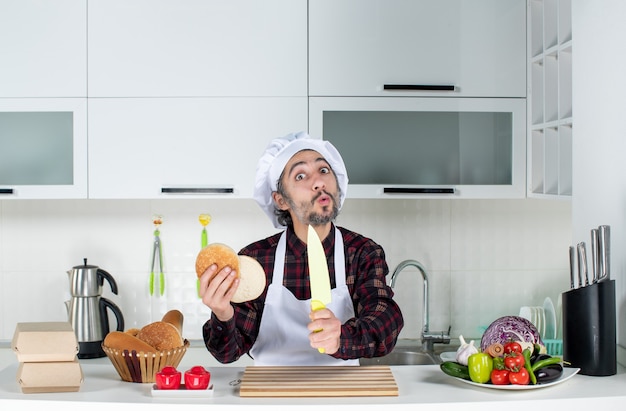 Vorderansicht des verwunderten männlichen kochs, der messer und brot in der küche hält