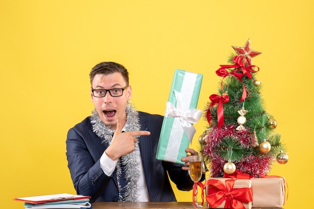 Vorderansicht des verwunderten Mannes, der auf Geschenk zeigt, das am Tisch nahe Weihnachtsbaum sitzt und auf Gelb präsentiert