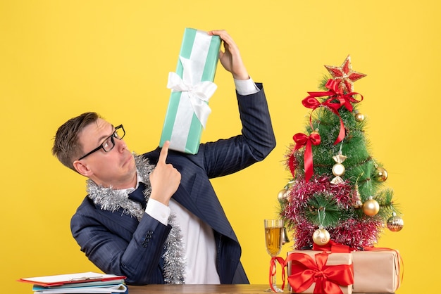 Vorderansicht des verwunderten Mannes, der Geschenk prüft, das am Tisch nahe Weihnachtsbaum und Geschenken auf Gelb sitzt