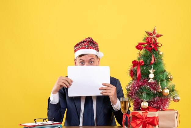 Vorderansicht des verwunderten Mannes, der Gesicht mit Dokumenten bedeckt, die am Tisch nahe Weihnachtsbaum sitzen und auf Gelb präsentieren