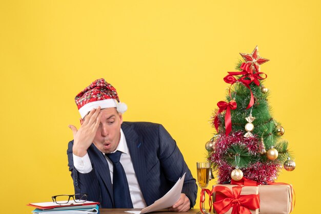 Vorderansicht des verwunderten Mannes, der Papiere prüft, die am Tisch nahe Weihnachtsbaum und Geschenken auf Gelb sitzen