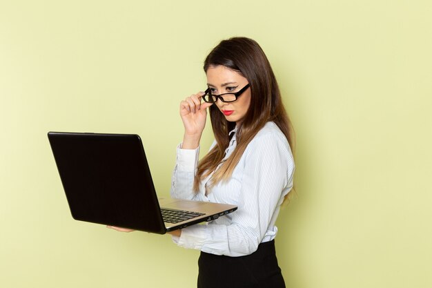 Vorderansicht des weiblichen Büroangestellten im weißen Hemd und im schwarzen Rock, der Laptop auf der hellgrünen Wand hält